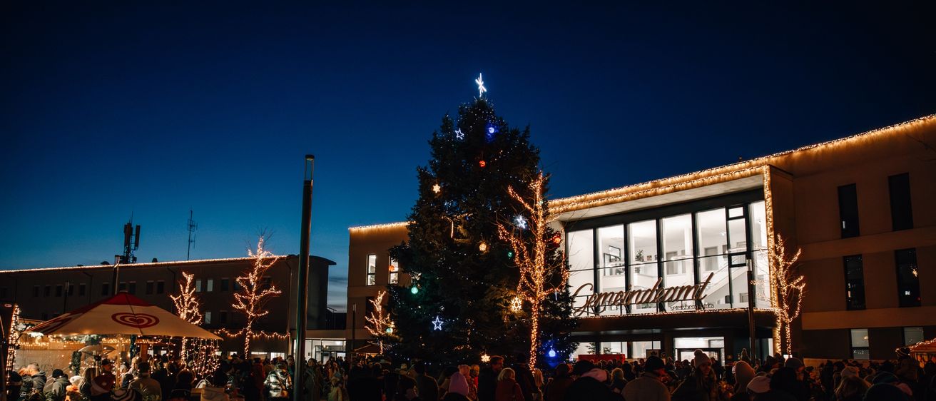 Eine Menschenmenge versammelt sich nachts um einen geschmückten Weihnachtsbaum. Der Baum ist mit Lichtern und Ornamenten verziert. Im Hintergrund ist ein Gebäude mit Glasfenstern und zusätzlichen Weihnachtslichtern zu sehen.