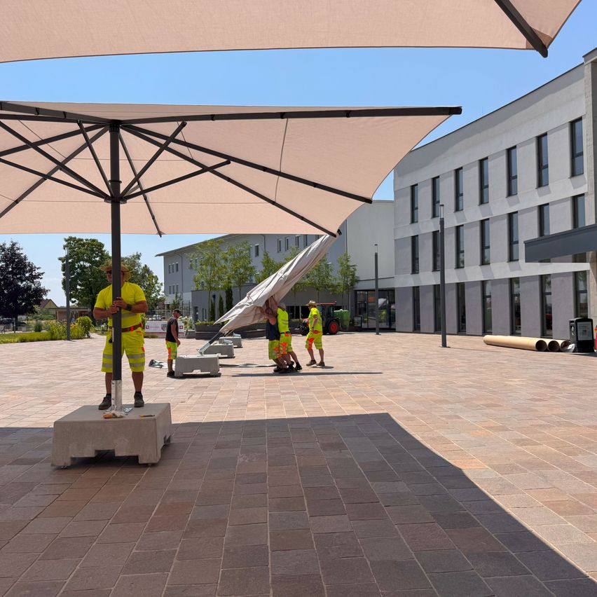 Eine Gruppe von Arbeitern installiert einen großen Schirm auf einer gefliesten Plaza vor einem Gebäude.