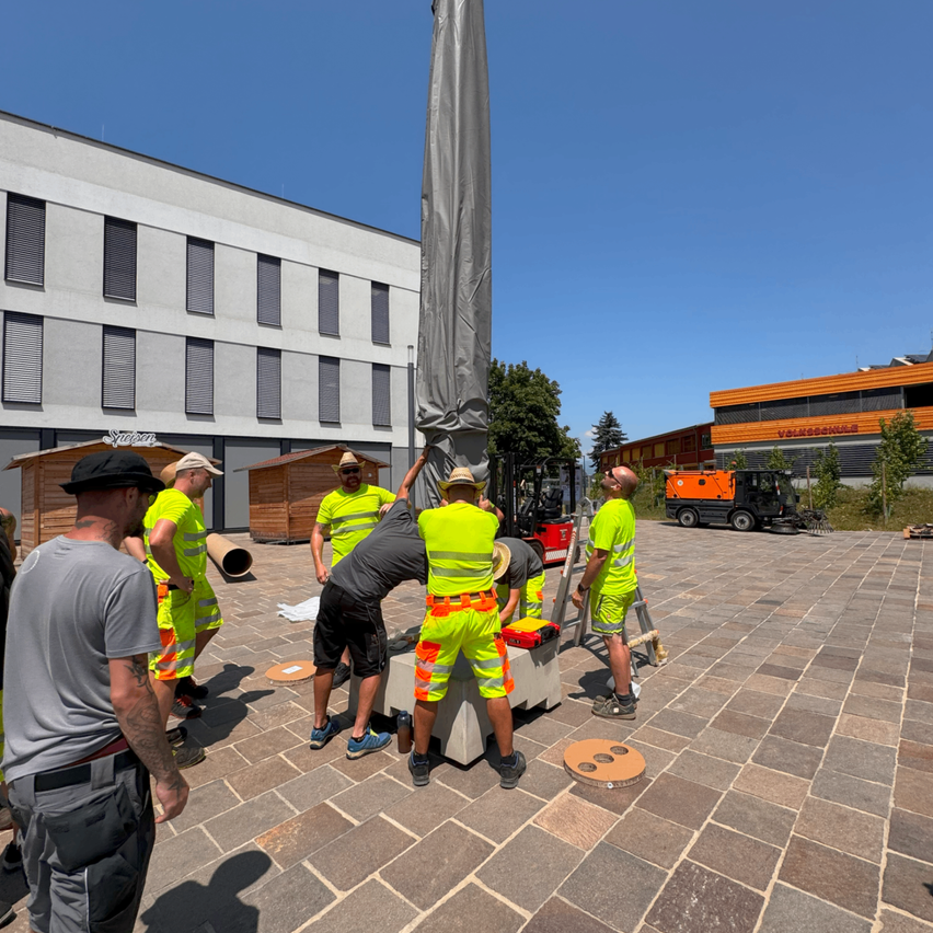 Eine Gruppe von Arbeitern in leuchtend grünen Westen richtet einen Pfosten vor einem Gebäude an einem sonnigen Tag auf.