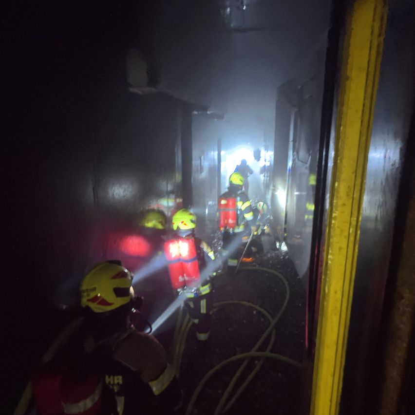 Feuerwehrleute in Schutzkleidung navigieren durch einen dunklen, rauchgefüllten Flur, einer hält eine Taschenlampe und ein anderer benutzt einen Schlauch.