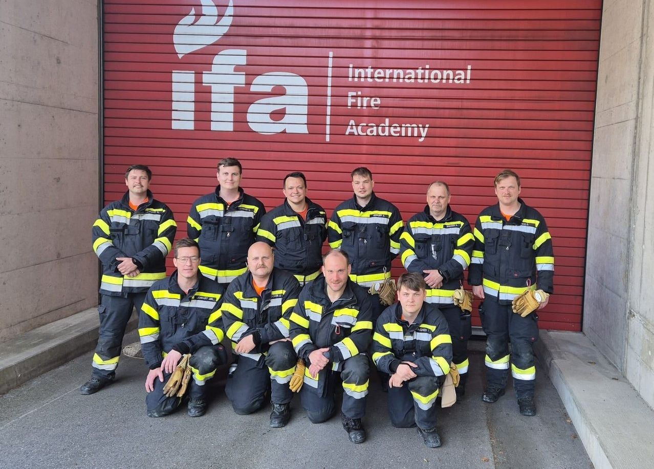Eine Gruppe von Feuerwehrleuten posiert vor der Internationalen Feuerwehrakademie, mit einem roten Gebäude und einem Logo mit einer Flamme im Hintergrund.