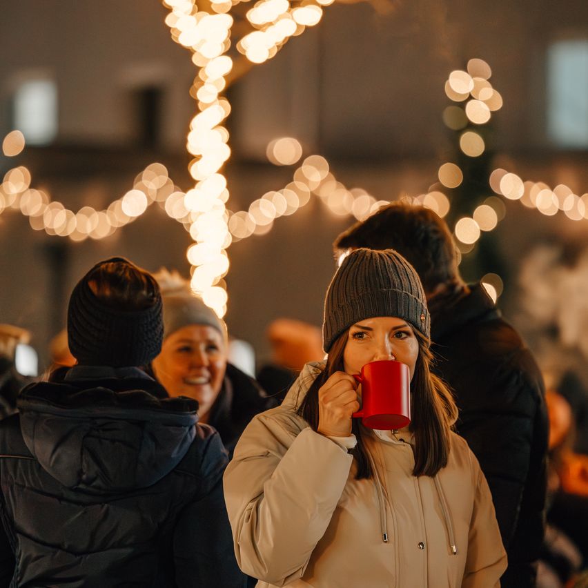 Eine Frau hält eine rote Tasse und lächelt, umgeben von Freunden bei einem festlichen Event unter leuchtenden Lichtern.