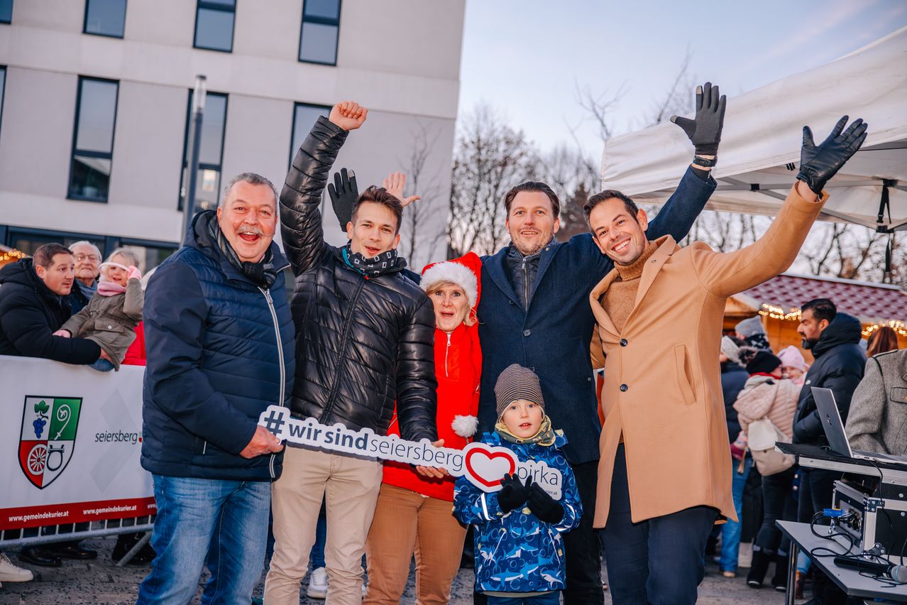 Eine Gruppe von Erwachsenen und einem Kind steht vor einem Gebäude, lächelt und posiert für ein Foto. Sie halten ein weißes Schild mit dem Hashtag #wirsindseisersberg. Dahinter befindet sich ein weißes Zelt.