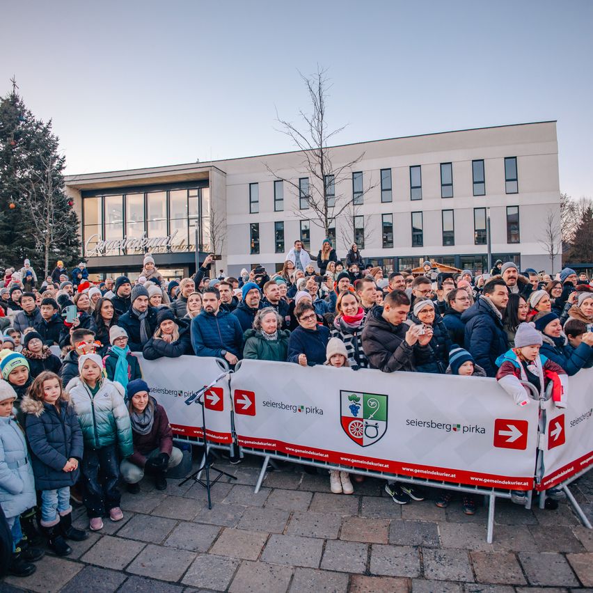 Eine große Menschenmenge versammelt sich vor einem modernen Gebäude, geschmückt mit Weihnachtsdekoration. Sie stehen vor einem roten Banner mit weißem Text. Einige Leute stehen auf Barrieren, während andere knien oder sitzen. In der Mitte ist ein Mikrofon aufgestellt.