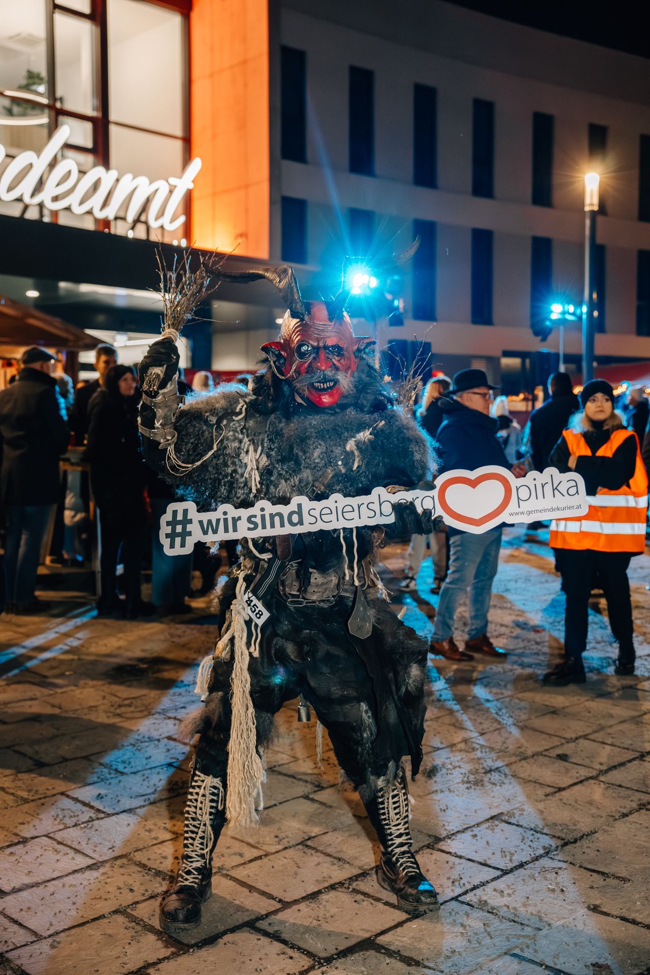 Eine Person in einem Kostüm mit Hörnern und einer Maske steht vor einer Menschenmenge. Er hält ein Schild mit der Aufschrift wir sind seiersborg. Eine Frau in einer Warnweste steht hinter ihm.