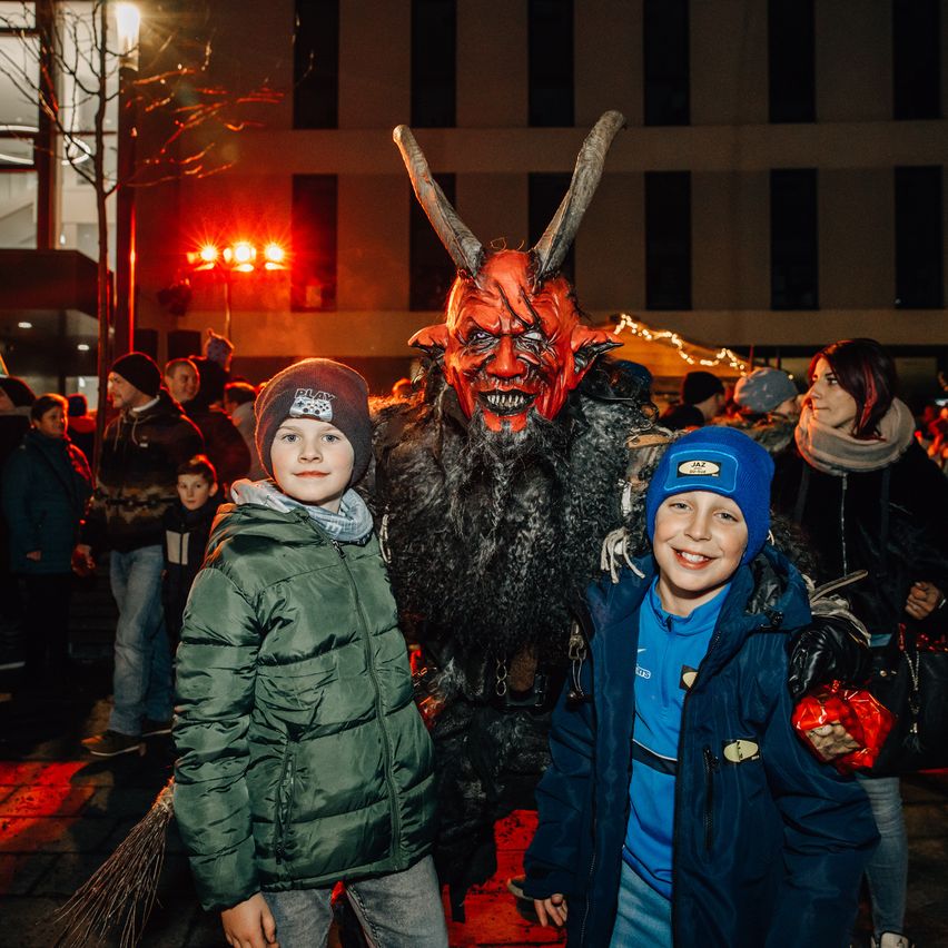 Zwei Kinder stehen neben einer als Teufel verkleideten Person, mit einer Menge im Hintergrund. Der Teufel trägt eine rote Maske mit Hörnern, und die Kinder tragen Winterkleidung.