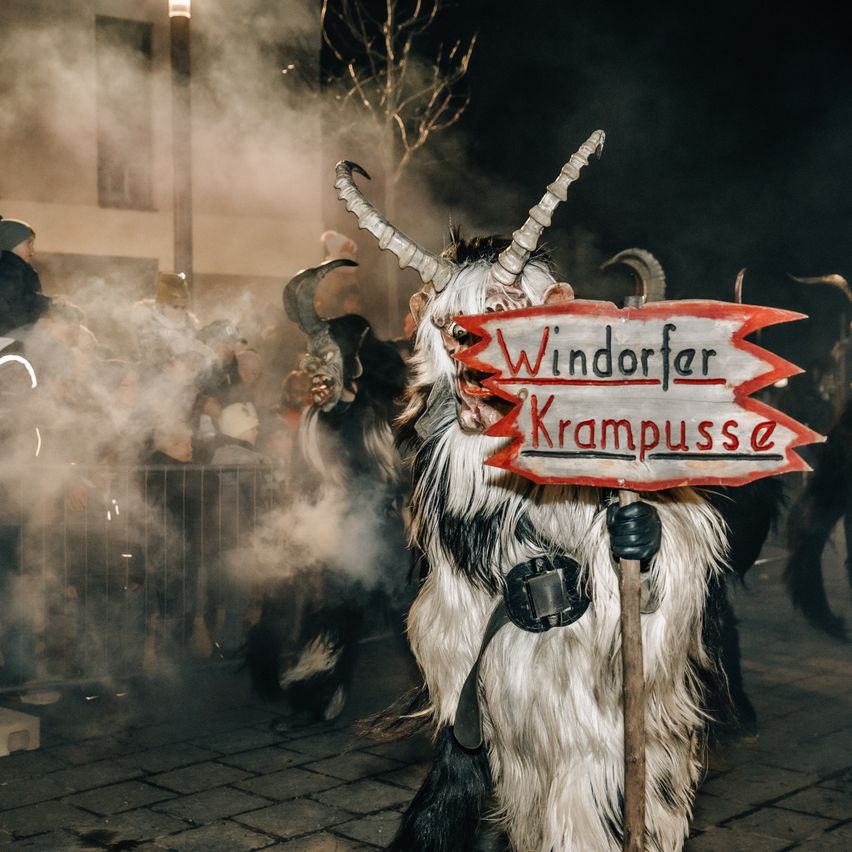 Eine Person, die als Krampus verkleidet ist und ein Schild mit den Worten Windopfer Krampus während einer Parade hält, mit Menschen, die hinter einem Zaun zusehen.