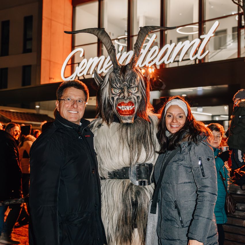 Ein Mann und eine Frau stehen zusammen mit einer Person in einem hörnertragenden Dämonenkostüm vor einem beleuchteten Gebäude bei Nacht.
