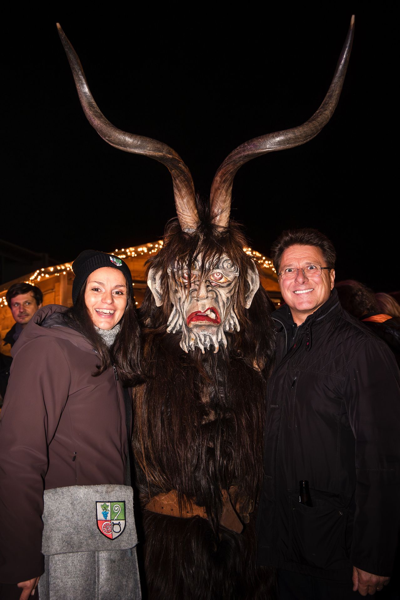 Ein Mann und eine Frau lächeln und posieren für ein Foto mit einer Person, die eine gehörnte, gruselige Maske trägt. Hinter ihnen sind einige Leute und beleuchtete Gebäude.