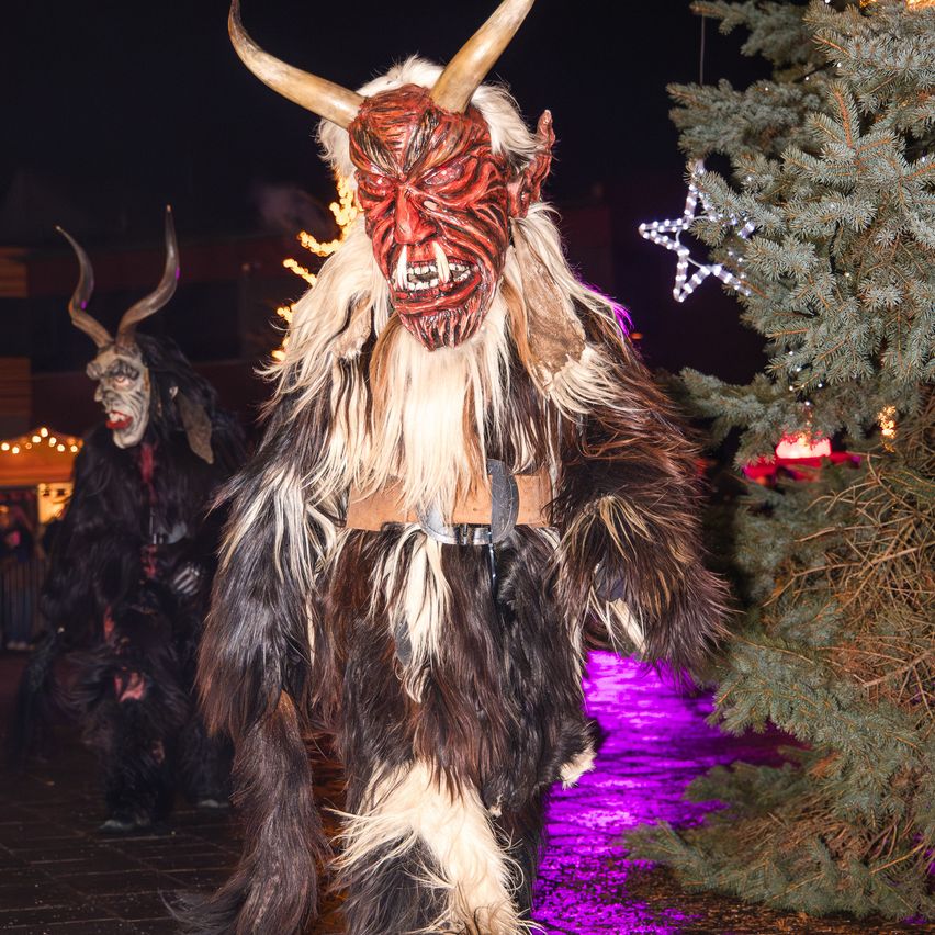Eine Person in einem gehörnten Kostüm geht neben einem geschmückten Weihnachtsbaum bei Nacht. Dahinter steht eine weitere Person in einem ähnlichen Kostüm. Die Szene ist mit lila Lichtern beleuchtet.