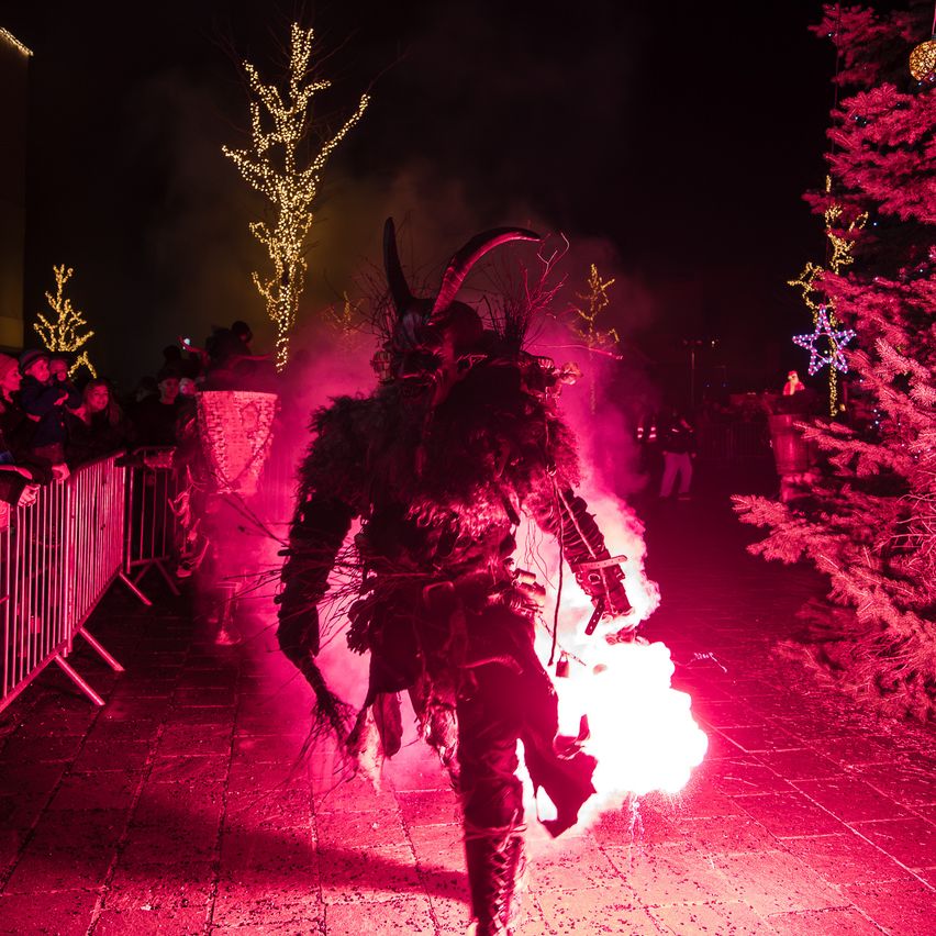 Ein Darsteller in einem gehörnten Kostüm geht durch eine Menge und erzeugt ein feuriges Spektakel auf einer rot beleuchteten Straße, umgeben von dekorierten Bäumen.