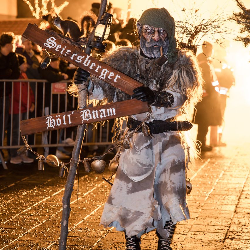 Eine Person in einem wilden Kostüm hält eine Laterne und zwei Holzschilder mit Text und Ketten in einem Außenbereich. Ein Feuer ist dahinter und eine Menge schaut zu.
