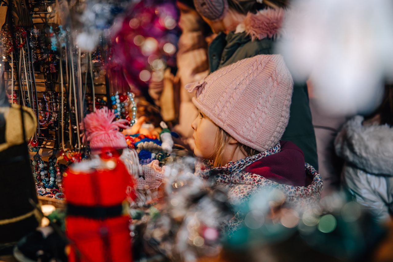 Ein kleines Mädchen mit einer rosa gestrickten Mütze betrachtet eine Verkaufsbude voller bunter Ornamente und Schmuckstücke auf einem festlichen Markt.