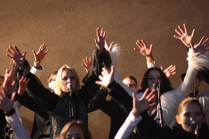 Eine Gruppe von Menschen, darunter zwei Frauen mit Mikrofonen, singt mit erhobenen Händen und sichtbarer Begeisterung.