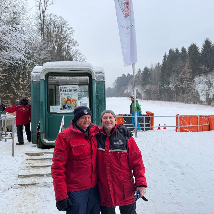 Zwei Männer in roten Jacken stehen nebeneinander im Schnee. Hinter ihnen ist eine kleine Hütte mit einem Plakat mit der Aufschrift 'Fasching' zu sehen. Ein Fahnenmast und ein verschneiter Hügel sind im Hintergrund.