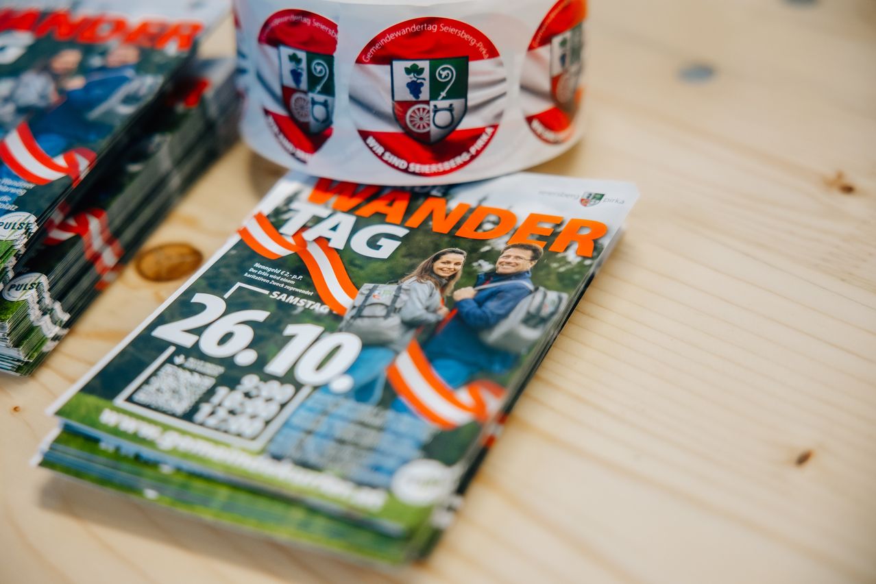 Ein Flyer für eine Wandertag-Veranstaltung am 26. Oktober liegt auf einem Holztisch. Eine Rolle mit Etiketten und einem Logo sowie einem Datum ist auf dem Flyer platziert.