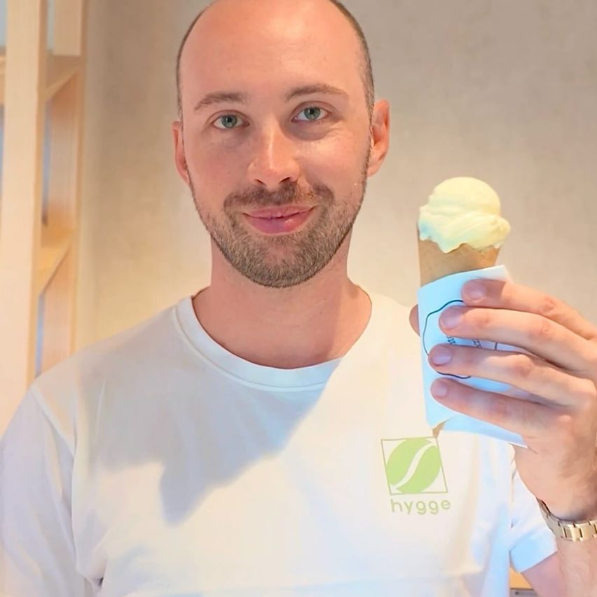 Ein Mann mit einem weißen T-Shirt mit einem Logo auf der linken Brust hält einen Eisbecher in der rechten Hand.
