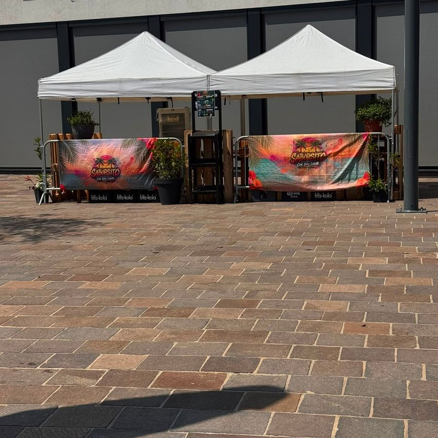 Zwei weiße Zelte mit bunten Bannern sind auf einem gepflasterten Weg aufgestellt. Jedes Zelt hat eine Holzbarriere und Topfpflanzen. Die Banner zeigen tropische Designs und den Text 'SABARITTO'.