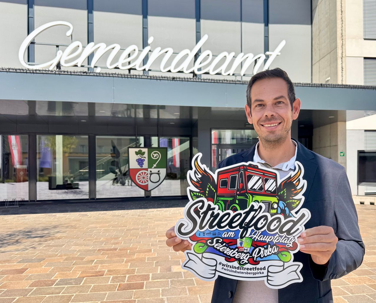 A man holds a colorful sign that says Streetfood in front of a modern building with a large sign that reads Gemeindeamt.