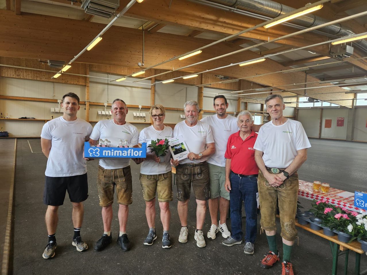 Sechs Männer in traditioneller bayerischer Kleidung stehen in einer großen Halle. Der Mann auf der linken Seite hält ein blaues Tablett mit einer Blume und einem Schild. Die anderen tragen weiße Hemden und Lederhosen. Sie scheinen für ein Foto zu posieren.
