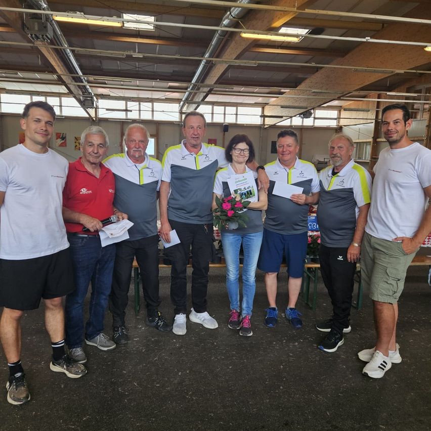 Eine Gruppe von Menschen in passenden Shirts und Sneakern steht in einer Halle, möglicherweise für ein Foto. Eine Frau hält einen Blumenstrauß und einige Männer halten Papiere.