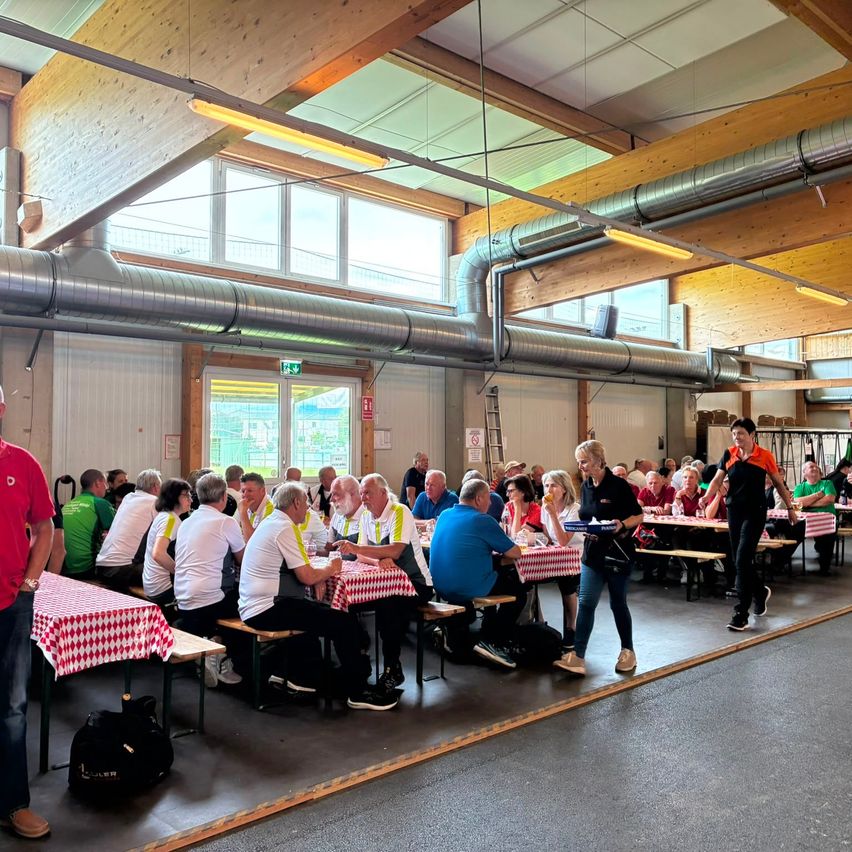 Eine Gruppe von Menschen diniert in einem großen Innenbereich mit Metallbalken und Fenstern. Einige sitzen an Tischen mit rot-weiß karierten Tischdecken, während andere stehen oder herumgehen.
