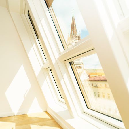 Ein Blick aus einem Zimmer mit Holzböden und weißen Wänden. Sonnenlicht strömt durch große Fenster, reflektiert sich auf dem Glas und zeigt einen Blick auf ein Gebäude mit einem Turm.