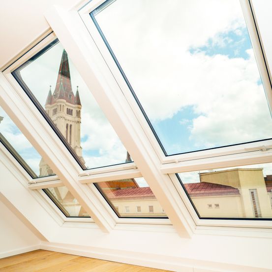 Ein Innenansicht eines Raumes mit weißen Wänden und Holzböden, ausgestattet mit drei großen, dreieckigen Fenstern, die einen Kirchturm und einen blauen Himmel mit Wolken reflektieren.
