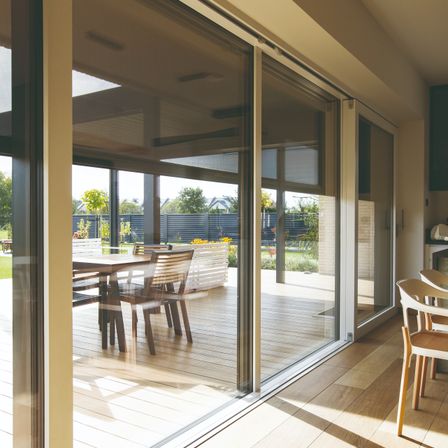 Ein Blick von innen in ein Haus, das eine Holzterrasse mit einem Esstisch und Stühlen, Schiebetüren aus Glas und einen Rasenbereich mit Bäumen und Pflanzen zeigt.