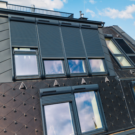 Ein modernes Gebäude mit geneigtem Dach hat Fenster mit grauen Rollläden. Das Äußere besteht aus Schindeln und hat einen Balkon oben.