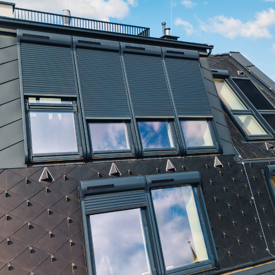 Ein modernes Gebäude mit geneigtem Dach hat Fenster mit grauen Rollläden. Das Äußere besteht aus Schindeln und hat einen Balkon oben.