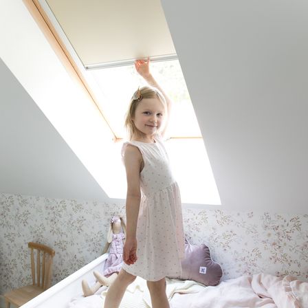 Ein junges Mädchen in einem Schlafzimmer steht neben einem Fenster, berührt die Jalousien, mit einer Puppe, die auf dem Bett liegt, einem Kissen und Blumentapeten.