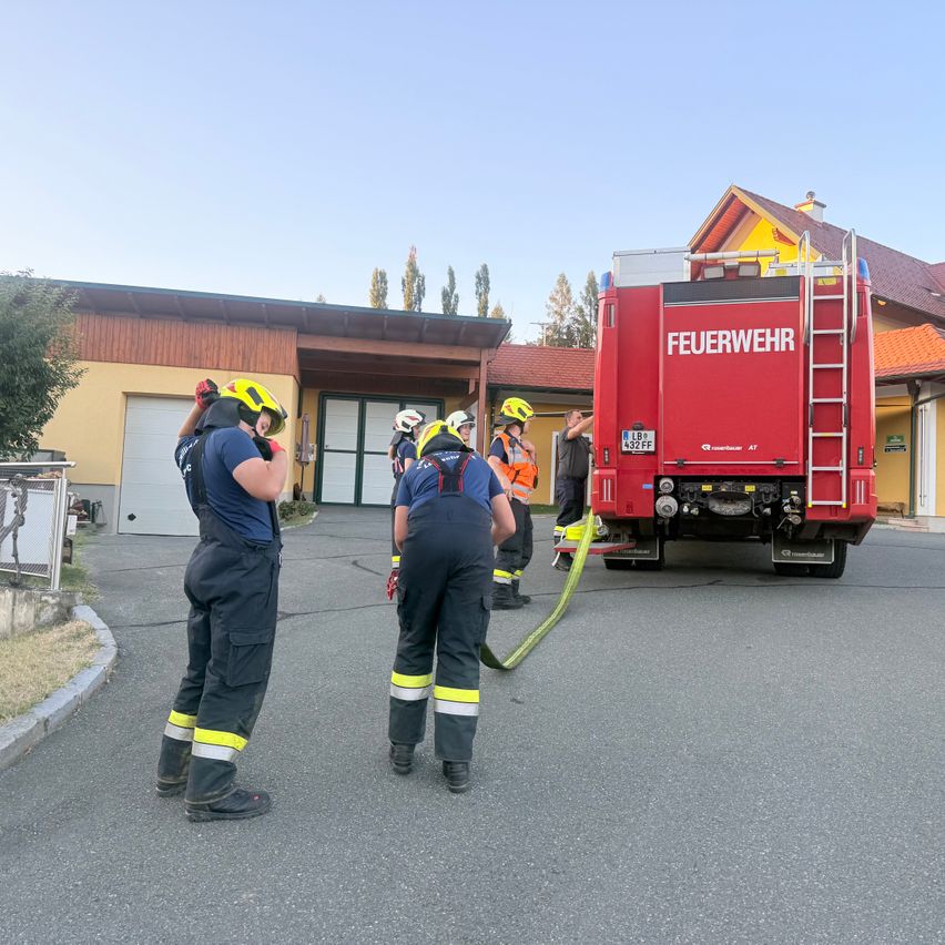 Firefighters in full gear are standing in front of a fire truck, with one holding a hose. They are in front of a building with a garage and a red roof.