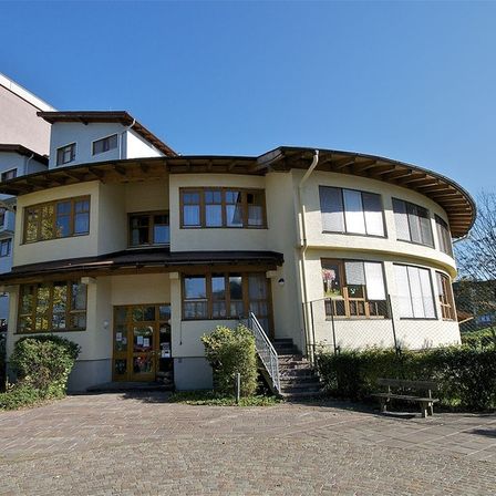 Ein gelbes, gebogenes Gebäude mit Glasfenstern und einer Treppe. Das Gebäude ist von einem Zaun und grünen Büschen umgeben.