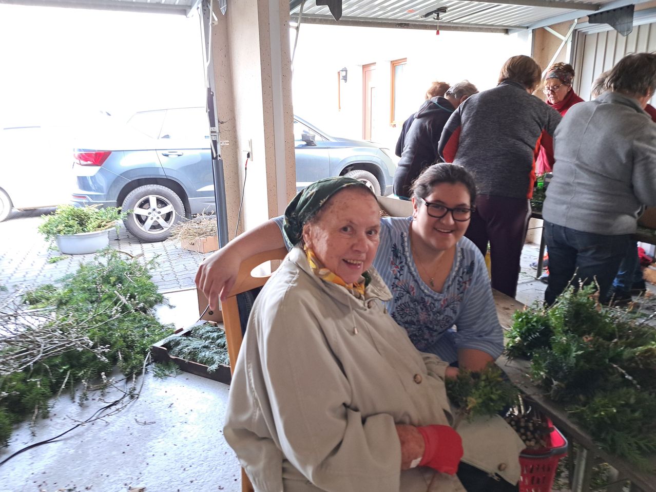Eine ältere Frau mit einem Schal sitzt auf einem Stuhl neben einer lächelnden Frau. Sie sind in einer Garage mit Pflanzen und Autos. Andere Leute stehen im Hintergrund.