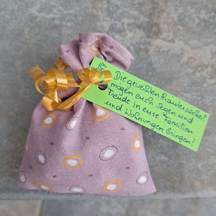 Ein kleiner lavendelfarbener Beutel mit einem gelben Band und einem grünen Anhänger mit handgeschriebenem Text auf Deutsch. Der Anhänger lautet 'Mögen die Wünsche wahr werden und dir Glück und ein neues Zuhause bringen!'.