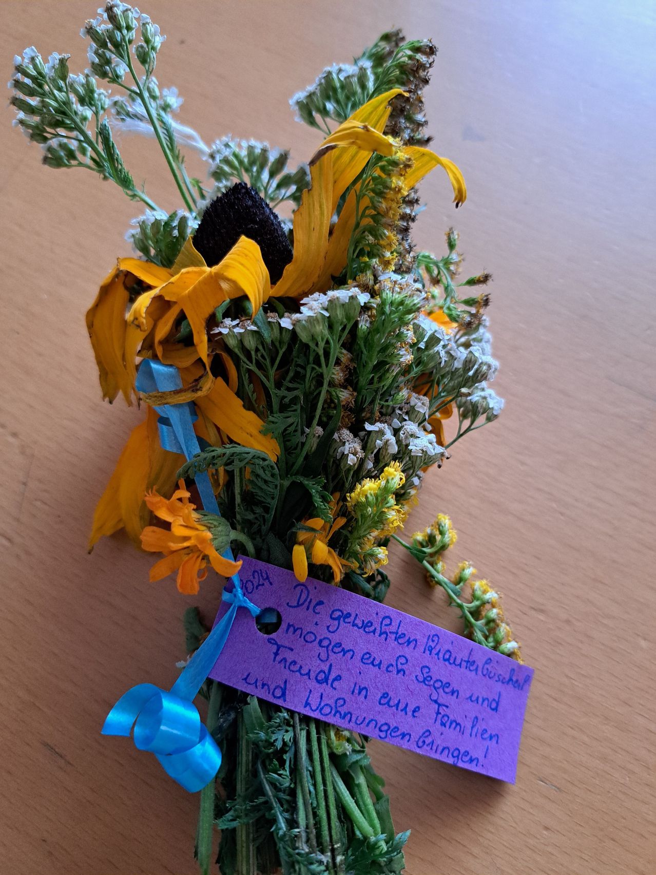 Ein kleiner Strauß getrockneter Blumen mit einem blauen Band und einem violetten Schild mit deutschem Text auf einer Holzoberfläche.
