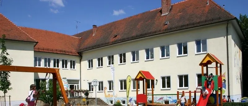Kinder spielen auf einem Spielplatz vor einem Gebäude mit roten Dächern. Es gibt Schaukeln und Rutschen. Einige Kinder klettern, während andere gehen.