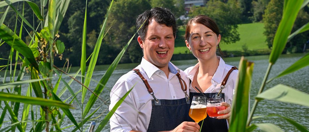 Ein lächelndes Paar steht auf einem Steg am See, jeder hält ein Glas Bier. Der Mann und die Frau sind in Jeansschürzen und weißen Hemden gekleidet, umgeben von hohen Pflanzen.