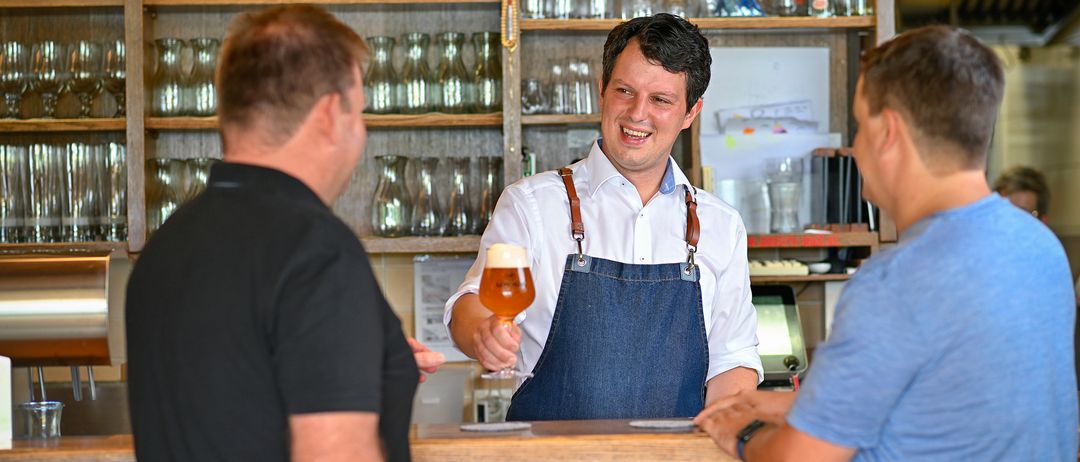 Ein Mann in einer Jeansschürze lächelt und hält ein Glas Bier hinter einer Bartheke. Er spricht mit zwei Männern vor ihm. Hinter ihm befinden sich Regale mit Gläsern und ein Halbmondschild.