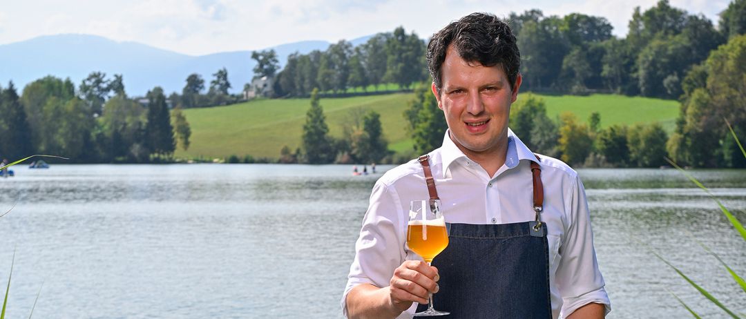 Ein Mann in einer Jeansschürze steht auf einem Steg, lächelt und hält ein Glas Bier in der Hand, vor einem See mit Bergen im Hintergrund.