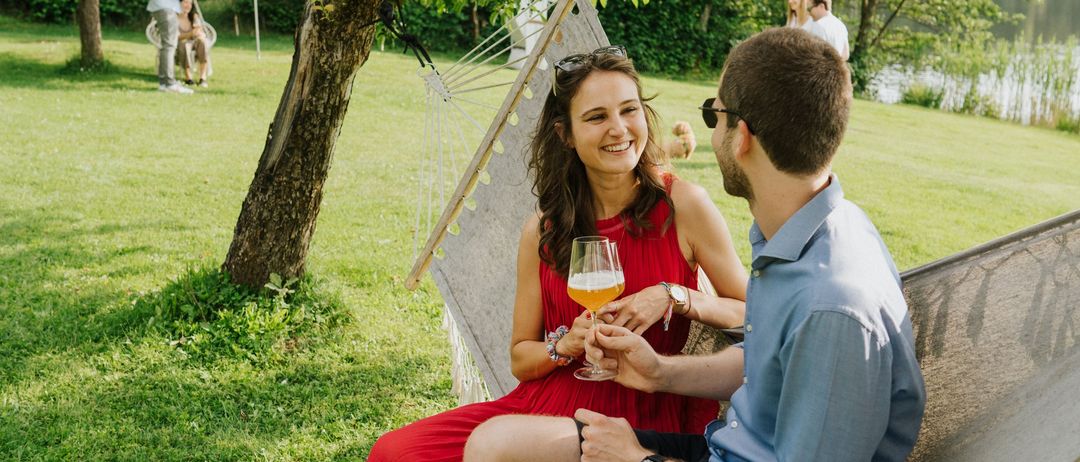 Ein Paar sitzt in einer Hängematte unter einem Baum und hält Gläser mit Bier. Ein See und andere Leute sind im Hintergrund.