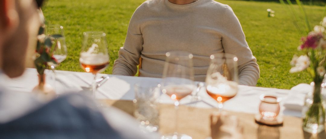 Zwei Männer sitzen an einem Tisch am See, mit Weingläsern und einer Blumenvase. Der Mann im Vordergrund trägt eine Sonnenbrille, während der Mann im Hintergrund lächelt.