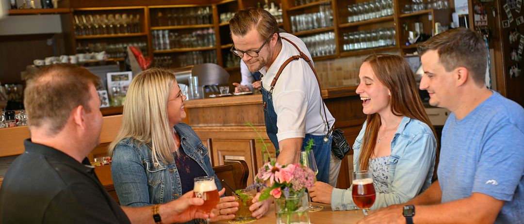 Eine Gruppe von Freunden genießt Getränke in einer Bar. Ein Mann steht hinter der Theke. Der Tisch ist mit Gläsern und einer Blumenvase gedeckt.