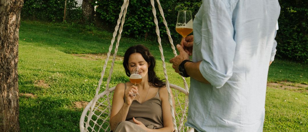 Ein Mann und eine Frau sind in einem Garten. Die Frau sitzt in einer weißen Hängematte und trinkt aus einem Glas. Der Mann steht neben ihr und hält ein Glas. Bäume und ein Haus sind hinter ihnen.