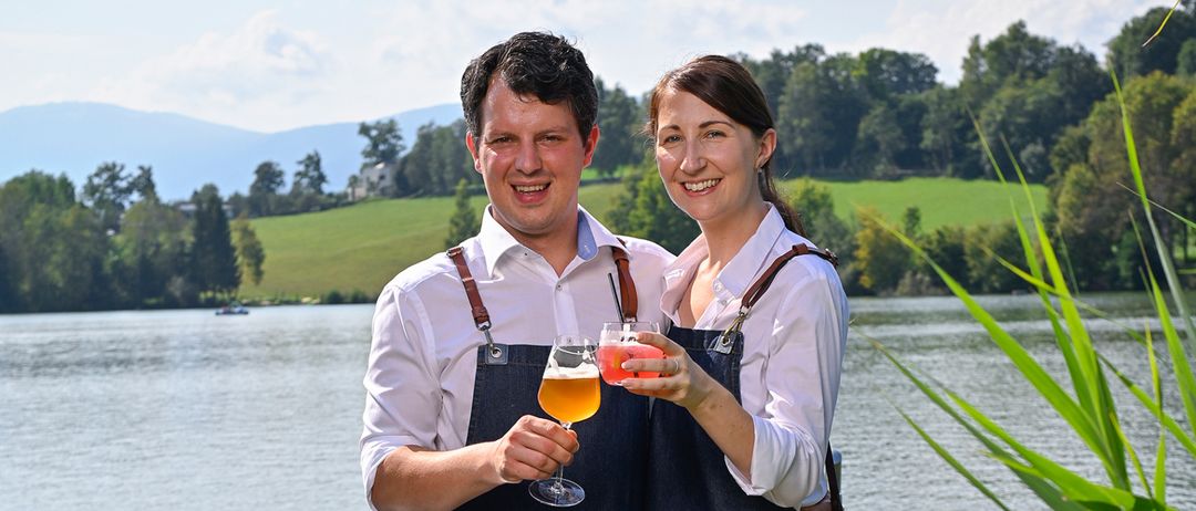 Ein Mann und eine Frau stehen auf einem Steg, lächeln und halten Getränke in Weingläsern. Sie tragen Jeansschürzen und Turnschuhe. Der See ist ruhig, mit einem Boot in der Ferne.