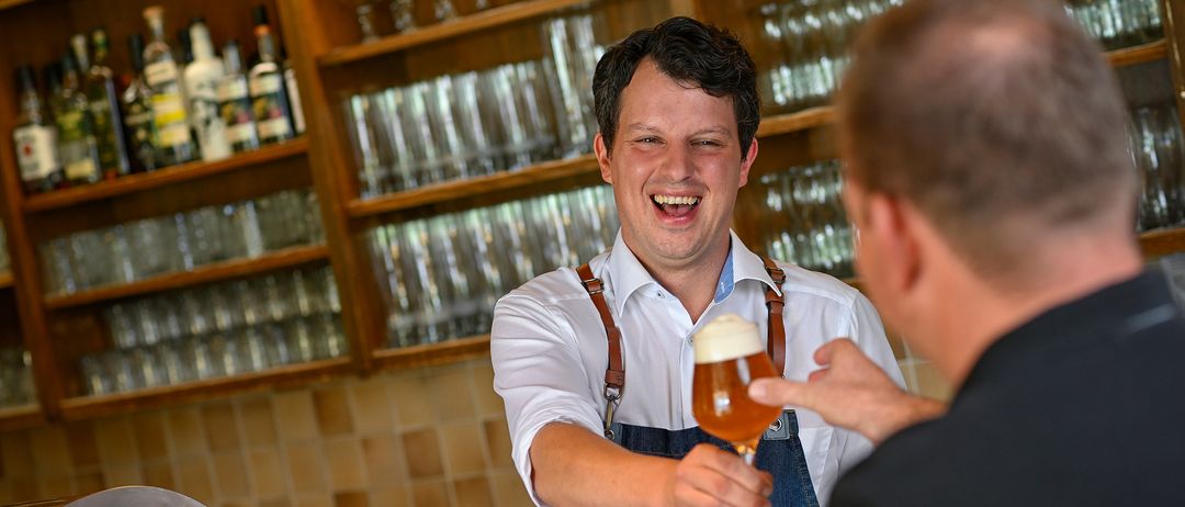 Ein Barkeeper in einer Schürze lächelt und reicht einem Kunden in einer Bar ein Glas Bier.