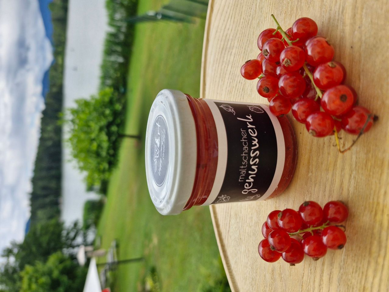 Ein Glas rote Johannisbeermarmelade steht auf einer Holzoberfläche mit roten Johannisbeeren darum. Das Glas hat einen weißen Deckel und ein schwarzes Etikett.