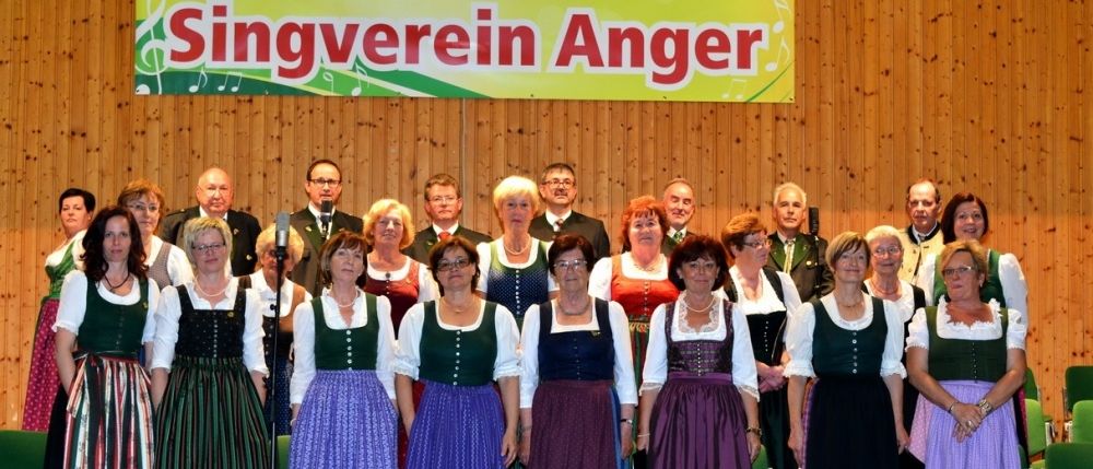 Eine Gruppe von Menschen in traditioneller deutscher Kleidung steht auf einer Bühne mit einem Banner, das 'Singverein Anger' im Hintergrund liest. Sie posieren für ein Foto.