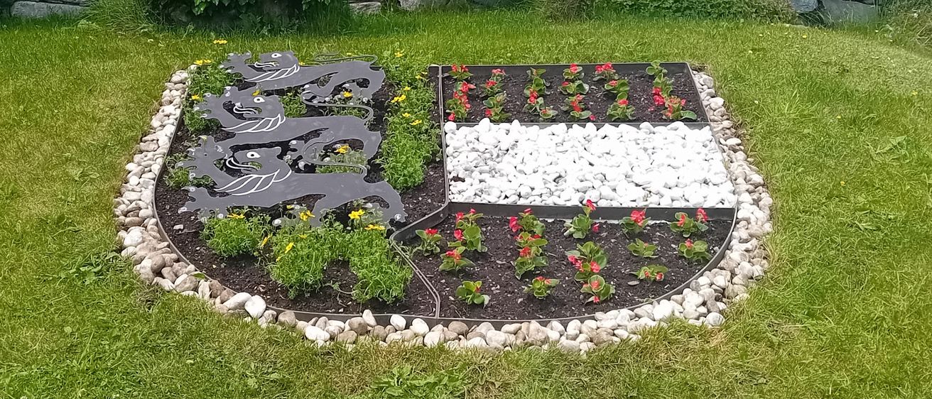 Garten mit einem zentralen Blumenbeet, umgeben von Steinen und Gras, mit einem Schild, das 'Karnten Blumende' beschriftet ist, mit einem Haus im Hintergrund, datiert am 5. Juni 2025 um 13:31 Uhr.